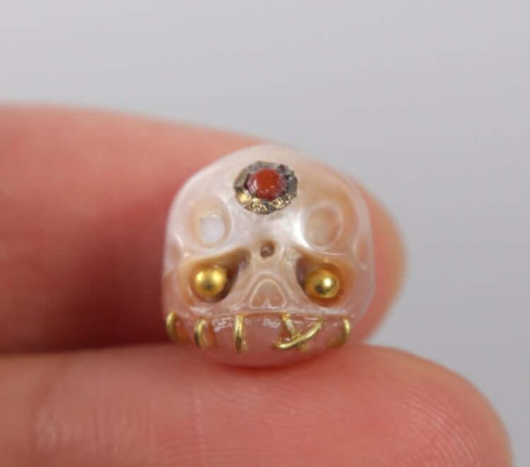 Hand holding a carved pearl skull bead with gold accents and a red gemstone detail, BelleJuri